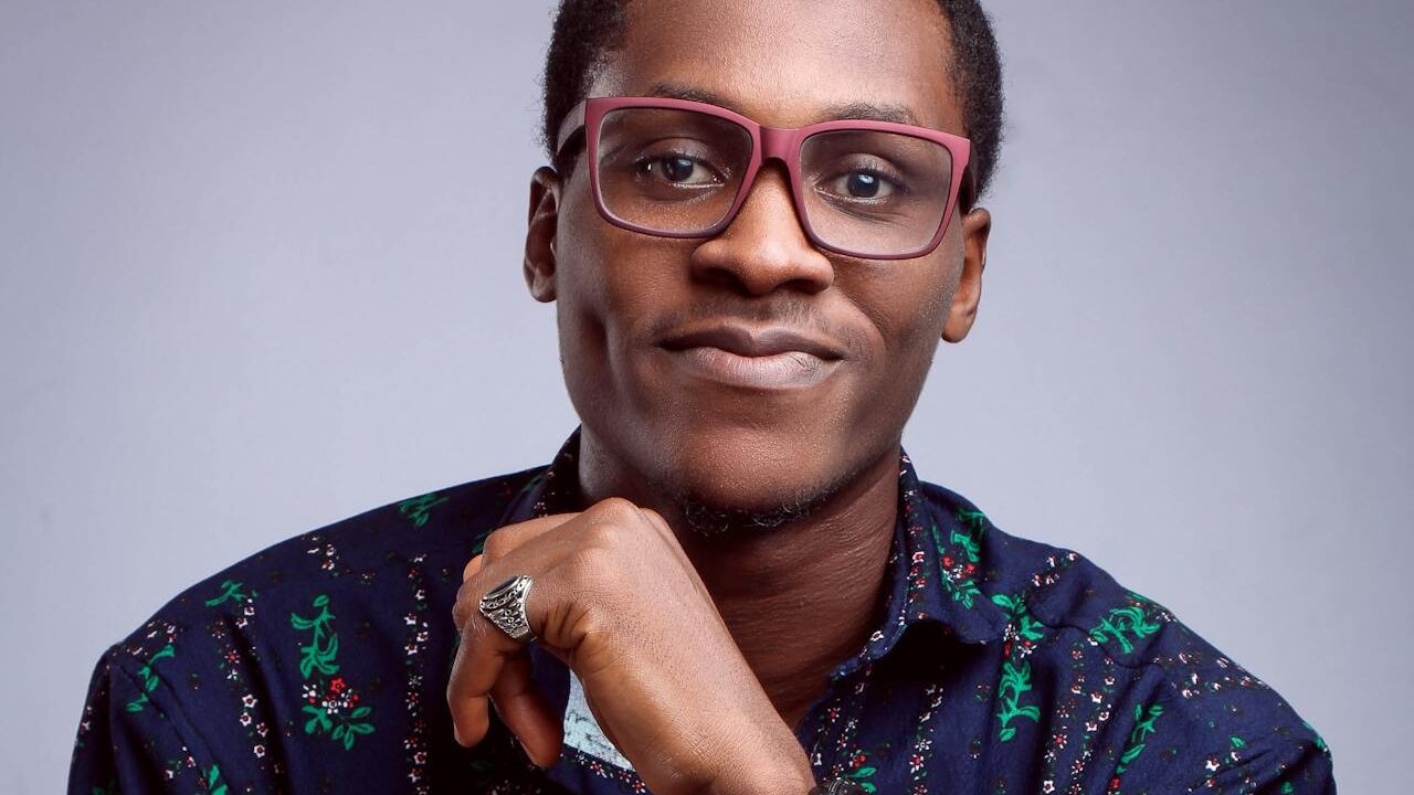 A stylish man with neatly trimmed hair, red glasses, and a floral-patterned shirt, posing thoughtfully with a ringed hand supporting his chin.