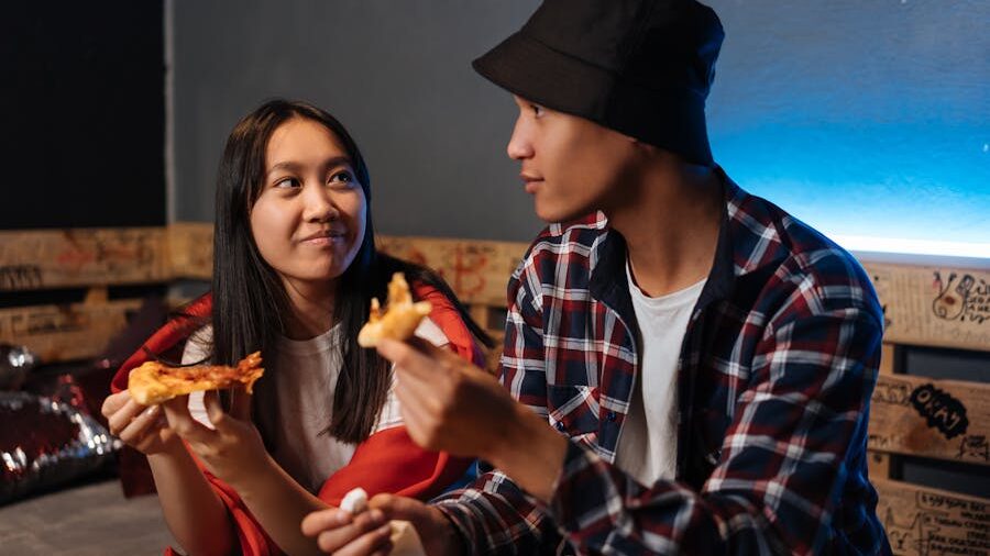 Two friends sharing a light moment while eating pizza, seated on wooden furniture with graffiti, under a blue light.