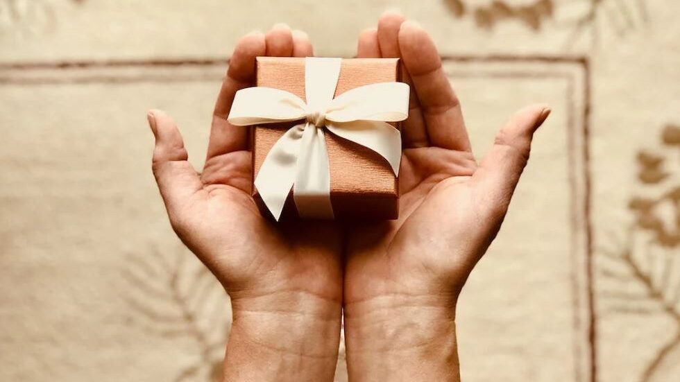 Hands holding a small gift box wrapped with a cream-colored ribbon, presented on a soft, beige background.