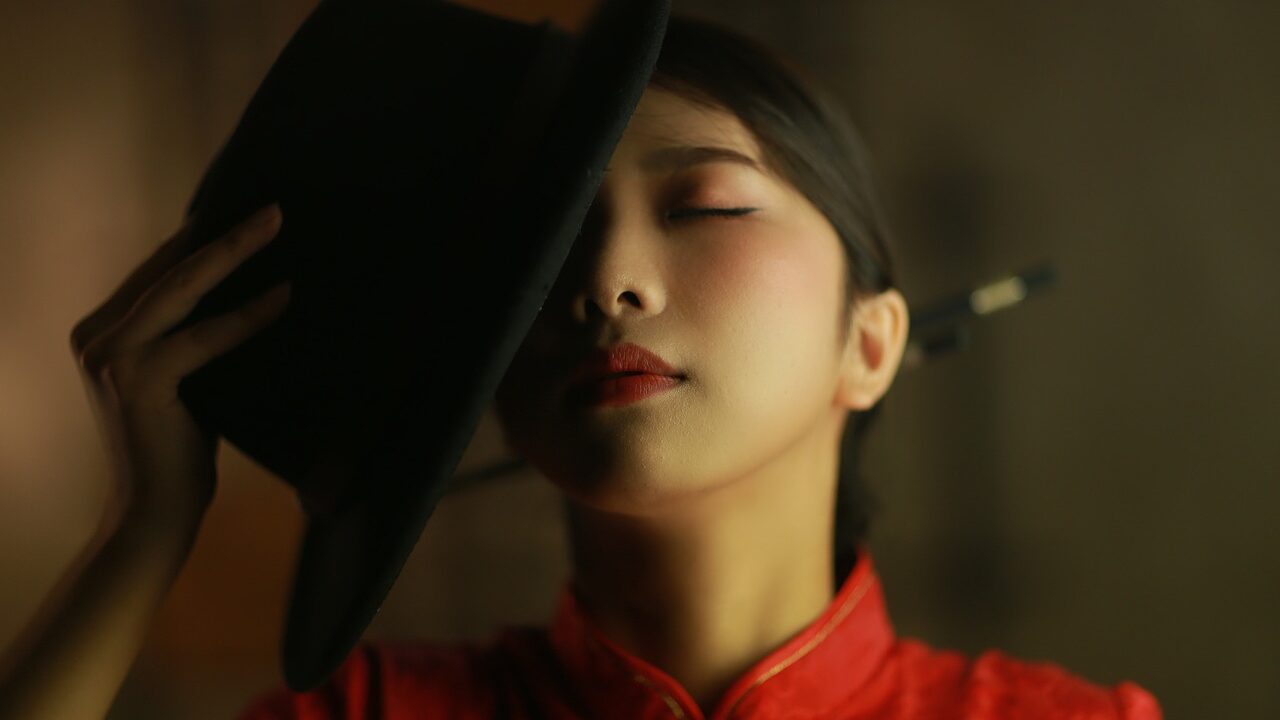 Individual in a red dress with elegant makeup, tilting her head slightly while holding a black hat in a dramatic pose.