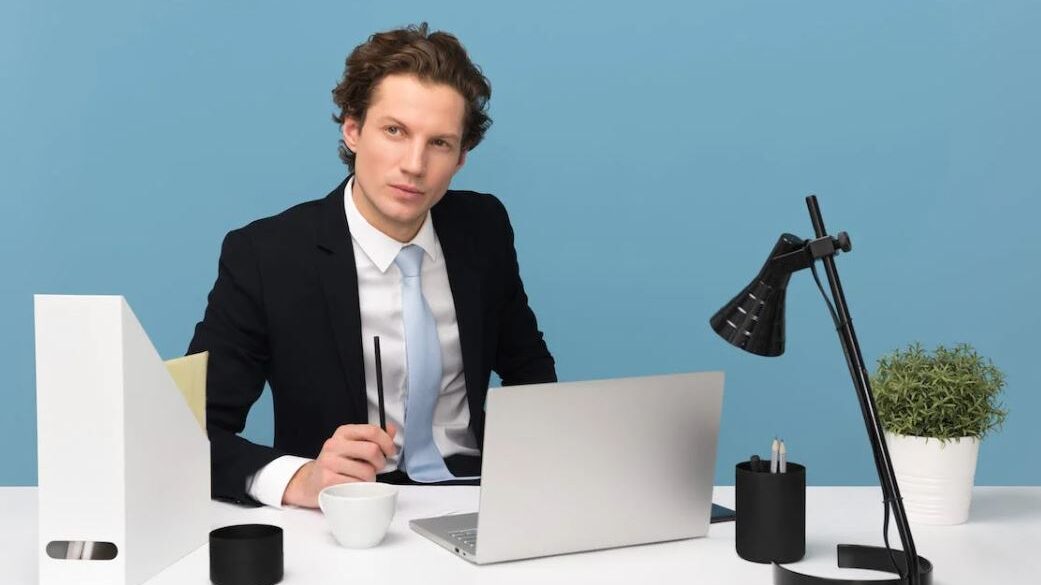 A young professional man with wavy brown hair wearing a suit, light blue tie, and holding a pen, sitting at a desk with a laptop, desk lamp, and potted plant, against a blue background.