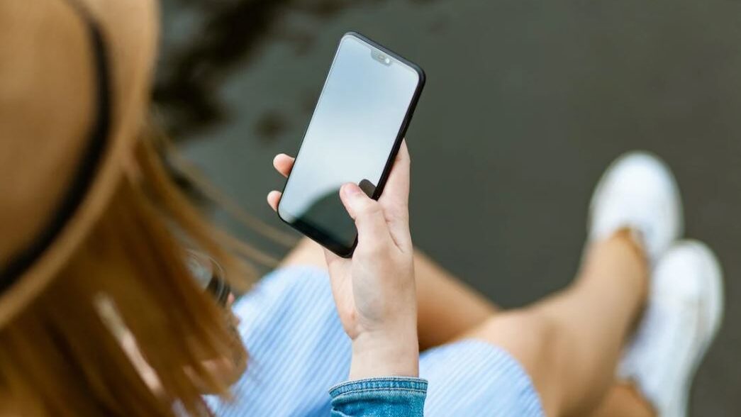 Individual sitting by the water, holding a phone and dressed casually, with focus on the smartphone.