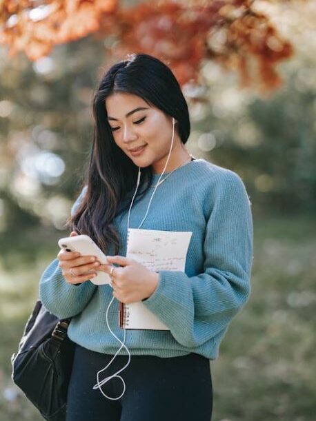 Young woman wearing headphones, looking at her phone while holding a notebook, standing outdoors in a park.