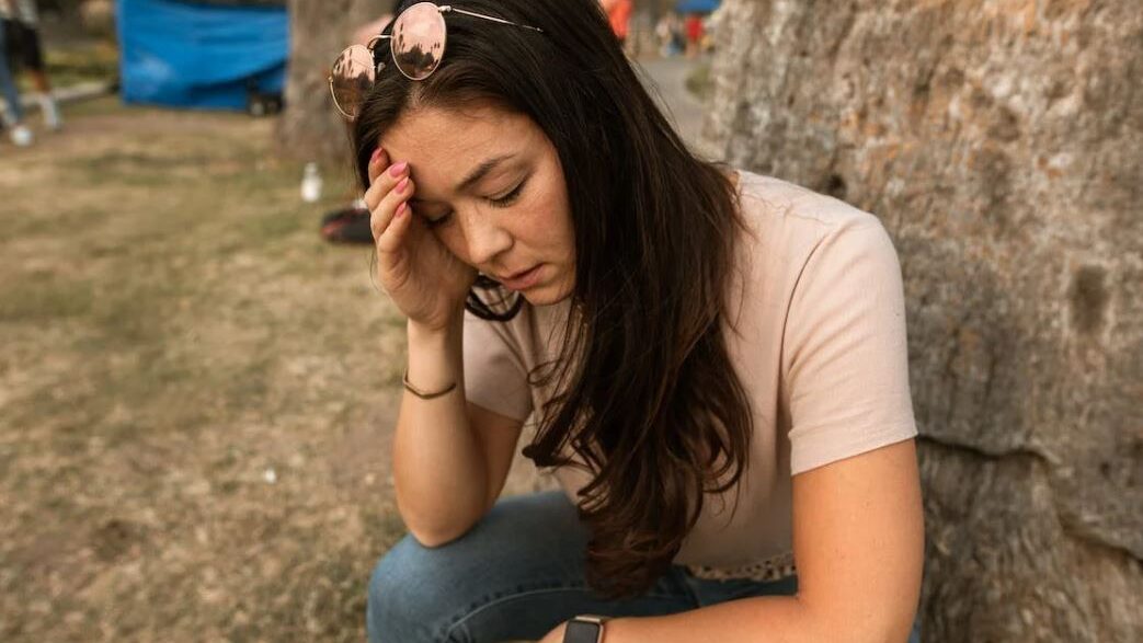 A young woman with a smartwatch and sunglasses, seated on the grass beside a rock, with a thoughtful expression in a park-like environment.