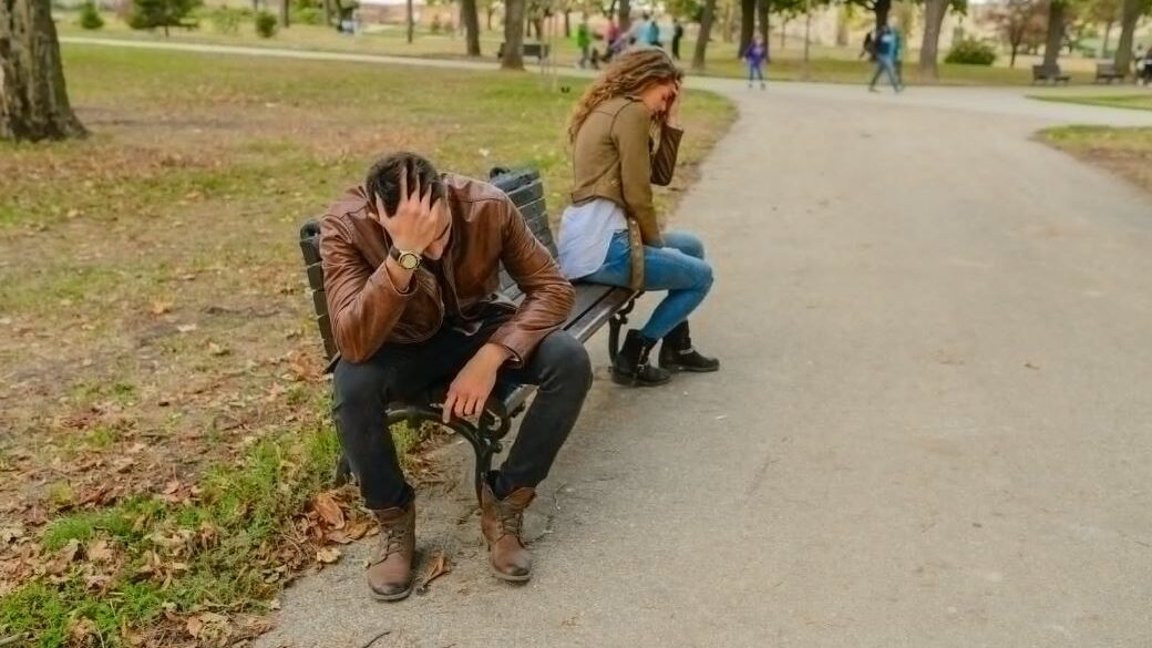 A couple in emotional conflict, sitting separately on a bench in an outdoor setting.