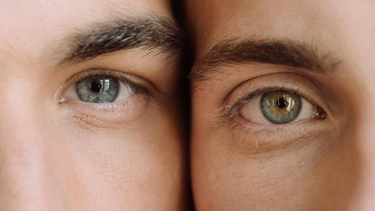 Two men’s eyes, one with a striking blue iris and the other with a hazel-green iris, framed by soft natural light and positioned symmetrically.