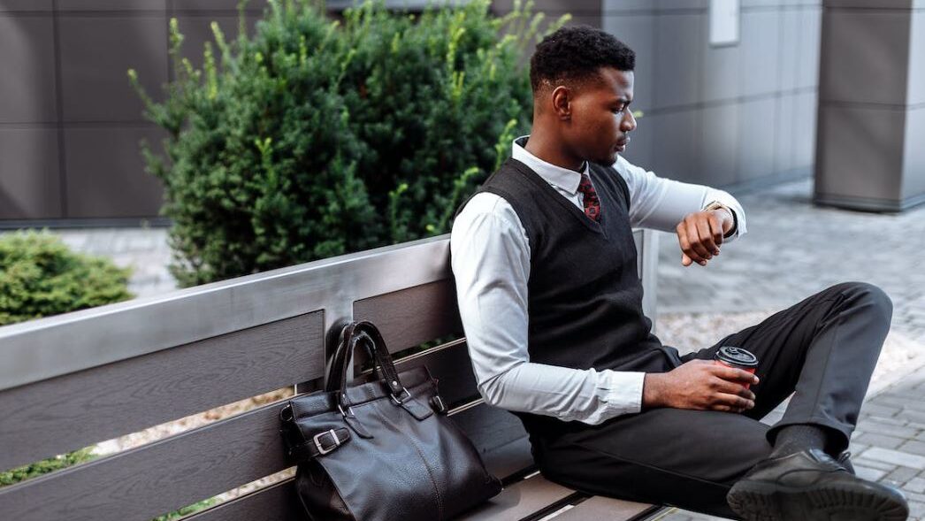 A man in business attire sitting on a bench, holding a coffee cup and checking his watch, with a leather bag placed beside him.