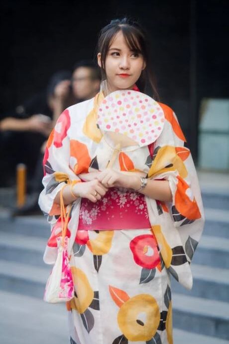 A woman dressed in a traditional Japanese yukata adorned with orange, yellow, and red floral designs, holding a small bag and a fan while gazing to the side.