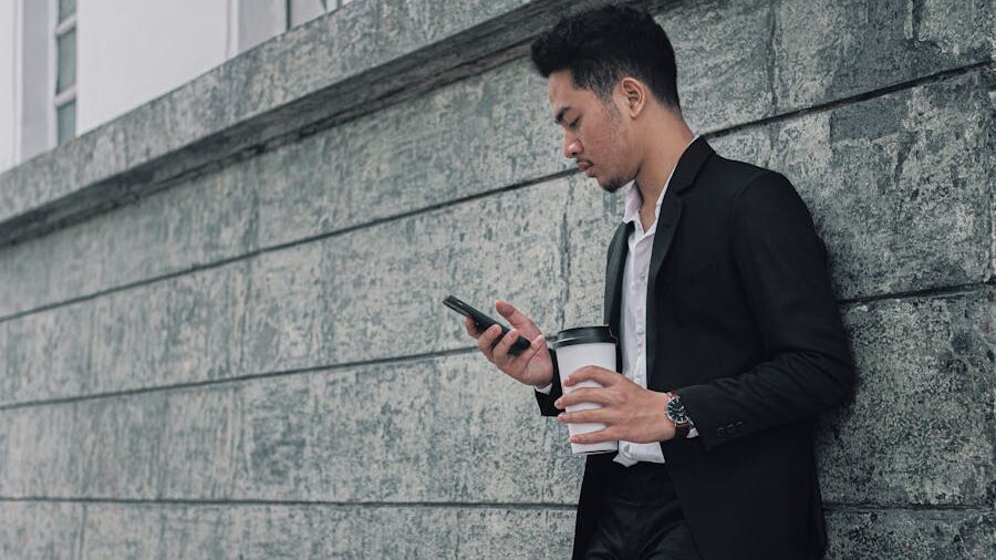 A man in a black suit leaning against a stone wall, holding a coffee cup and checking his phone.