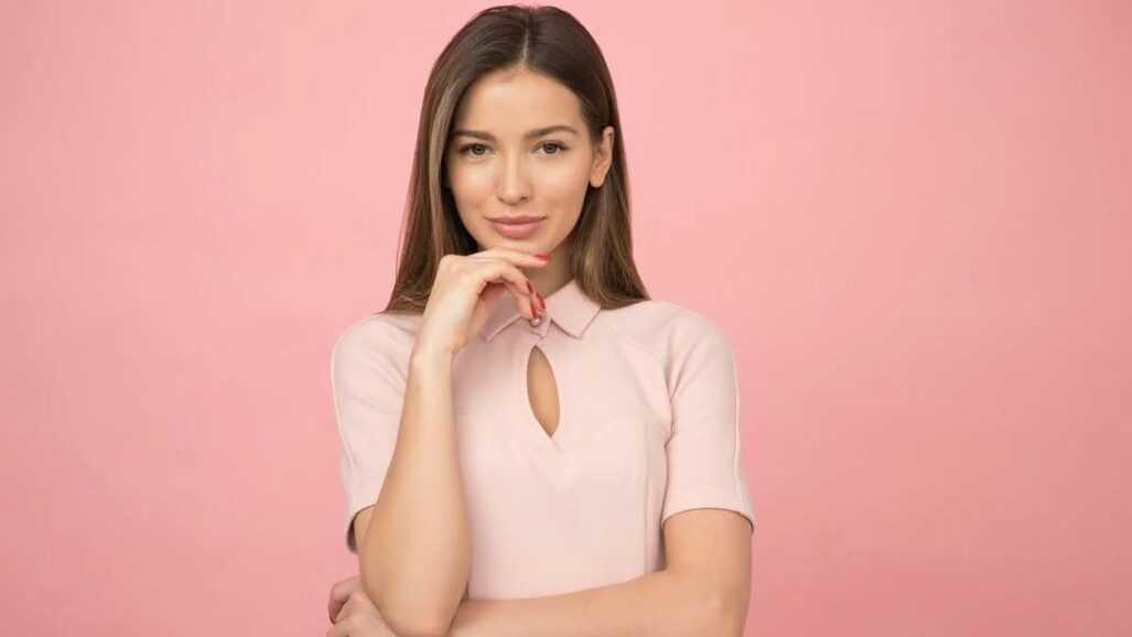 A woman wearing a pale pink top, resting her chin on her hand, with a soft smile against a pink background.