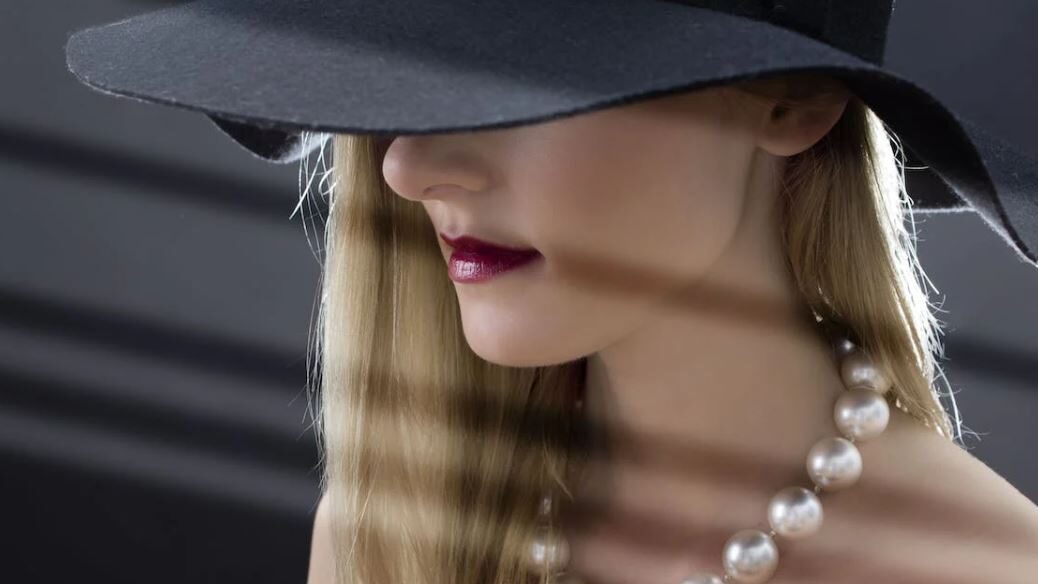 A woman wearing a wide-brimmed black hat and pearl necklace, partially obscured by shadow.