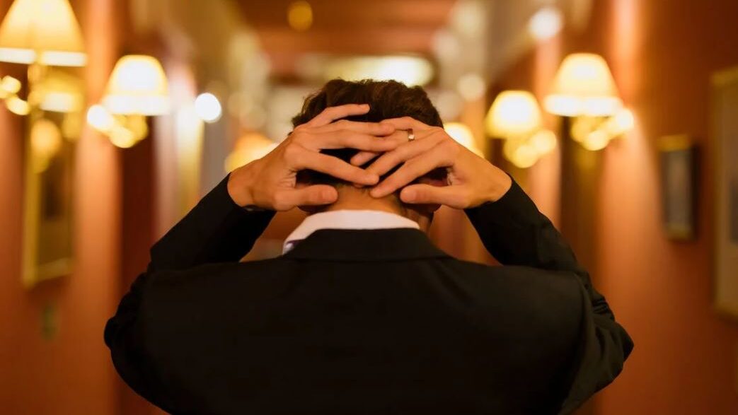 A man in formal attire with hands resting on his head in an elegant hallway.