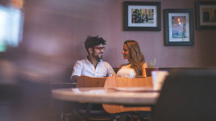 Couple sitting at a table in a cafe, looking at each other with affectionate smiles, capturing a candid moment of connection.