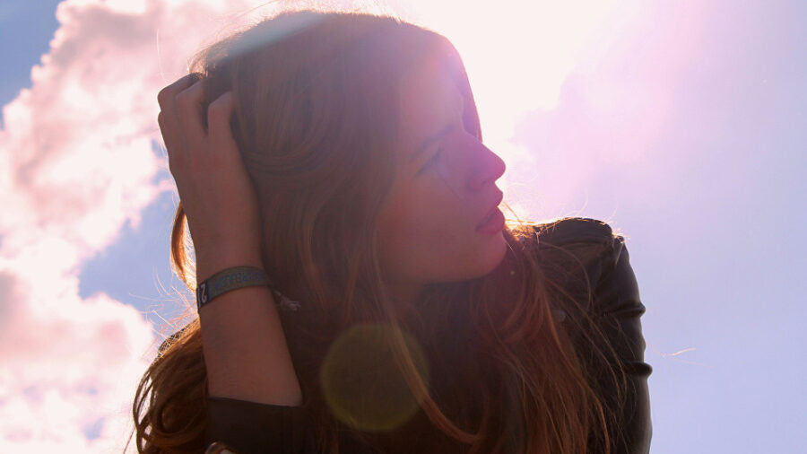 Backlit portrait of a woman touching her hair, with sunlight and clouds creating a soft, dreamy atmosphere.