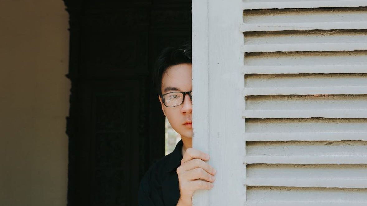 A person wearing glasses peeking from behind a wooden shutter, with half of their face visible.