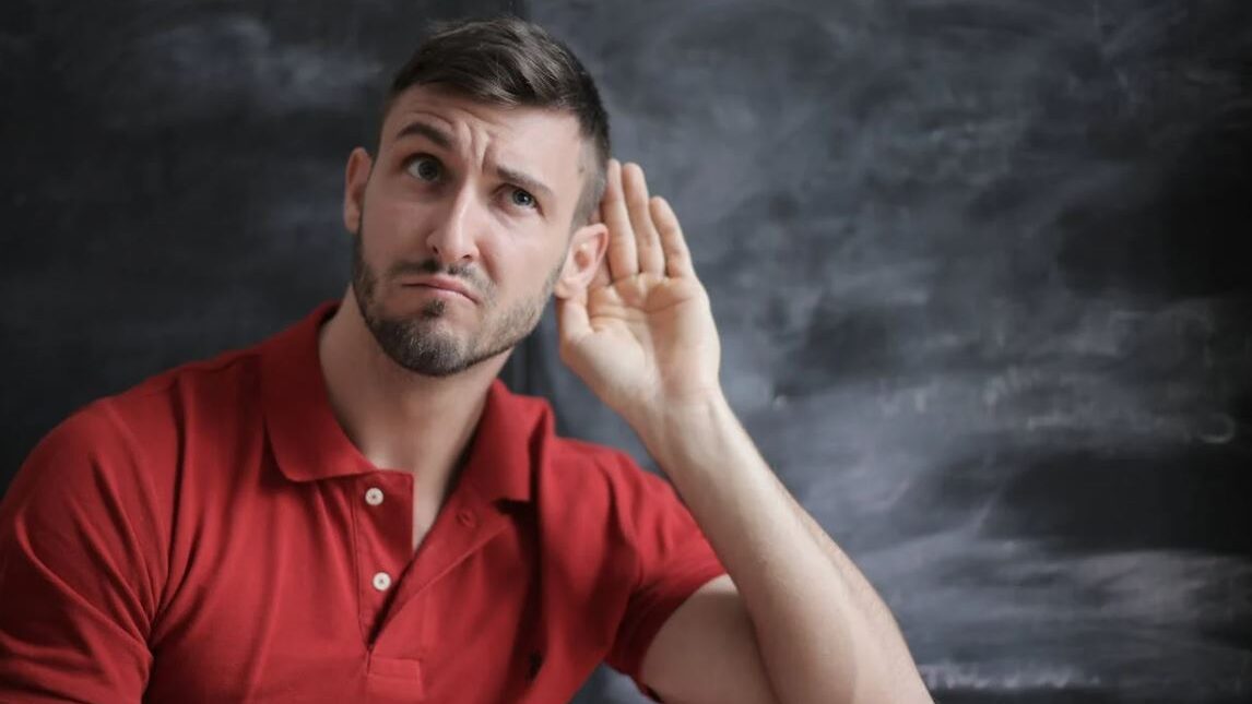 A man with a beard and short hair, gesturing to listen more closely, against a blackboard backdrop.