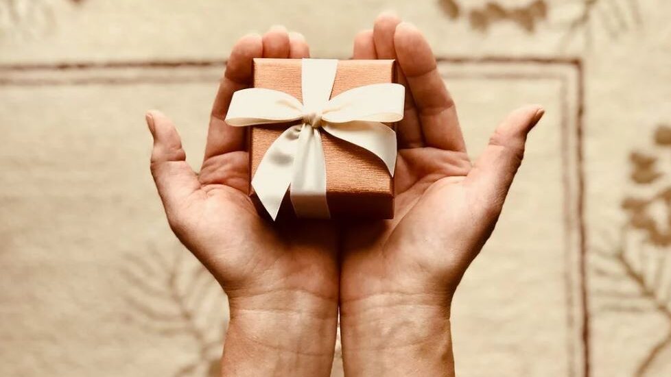 Hands holding a small gift box with a white ribbon, set against a soft, patterned background.