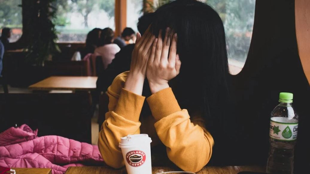 A person hiding their face with their hands in a coffee shop, a drink and a water bottle on the table.