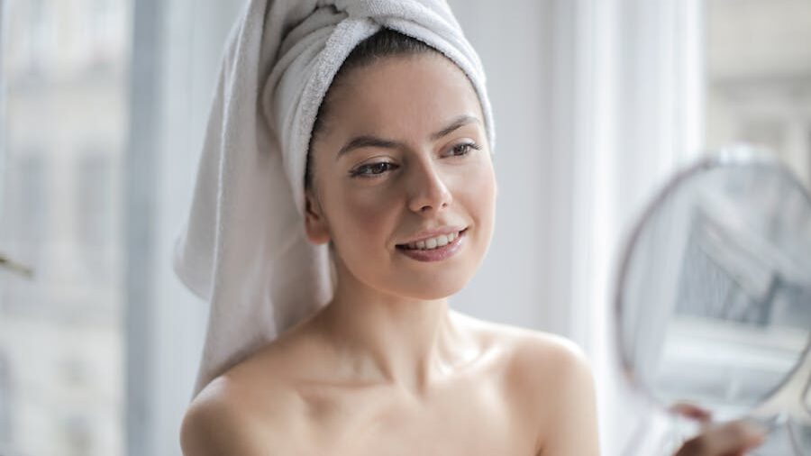 A woman with a towel wrapped around her head smiling into a handheld mirror near a bright window.