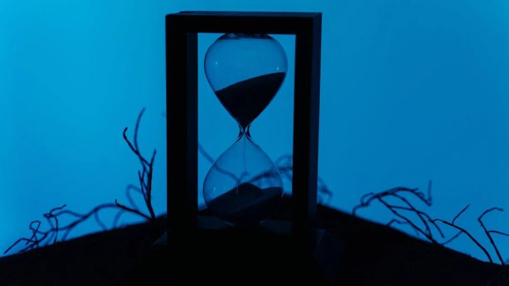 An hourglass in a rectangular frame, placed against a blue background with shadowed branches.