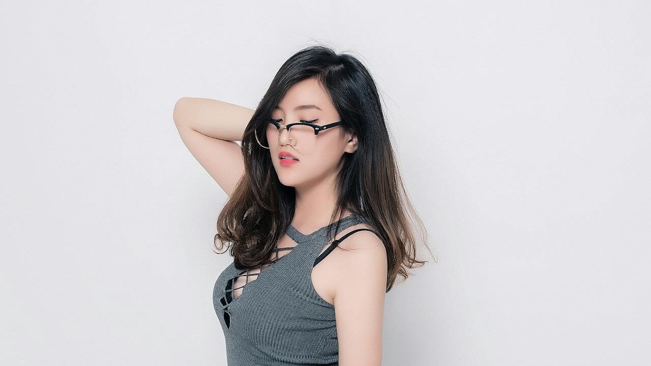 Confident young woman in a stylish gray dress poses with one hand on her head, adjusting her glasses with a bold expression.