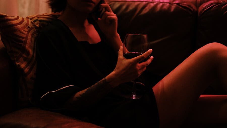 A relaxed person sitting on a leather couch in moody dim lighting, holding a glass of red wine while engaged in a phone conversation.