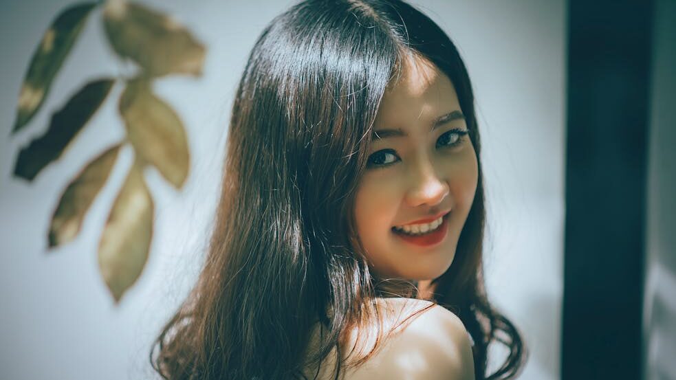 A young trans woman with long dark hair smiling over her shoulder, in soft natural light, with a decorative golden leaf in the background.