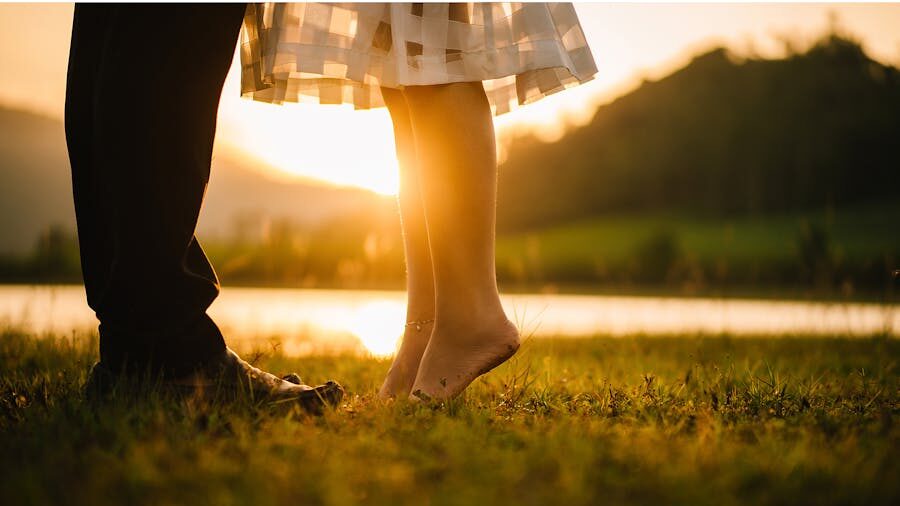 A romantic moment captured at sunset, showing a barefoot person on tiptoes reaching up to kiss their partner near a lake.