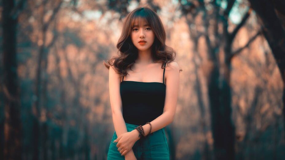 A young trans woman with long wavy hair standing in a forest, wearing a black top and green pants, with a calm expression.