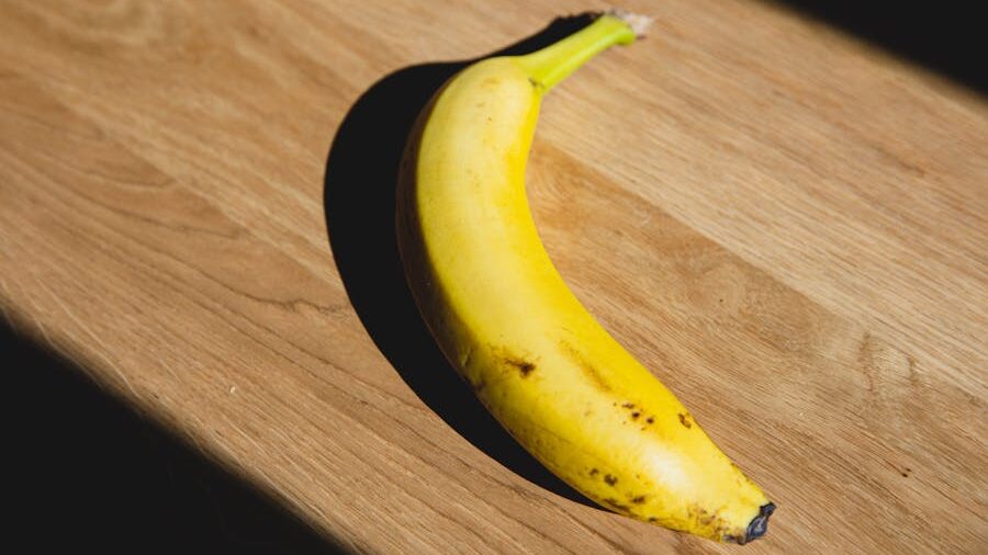 A single yellow banana with a few brown spots, placed on a wooden surface in natural light.