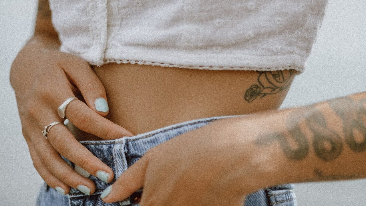 Cropped view of a waist adorned with a small floral tattoo, pale blue nail polish, and casual denim.