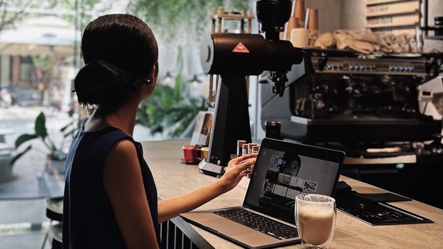  Woman in a dark blue dress sitting at a café counter, working on a laptop with a coffee machine in the background.