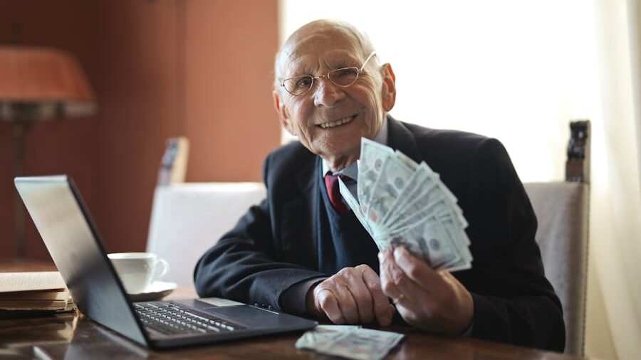 Elderly man in a suit and glasses sitting at a desk, holding a fan of dollar bills with a laptop in front of him.