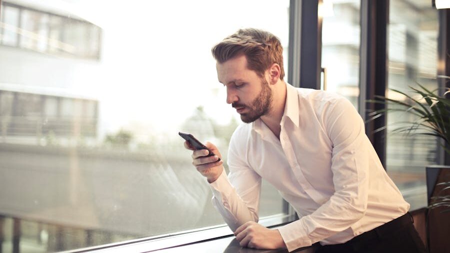 Bearded man in business attire checking his phone by a large window.