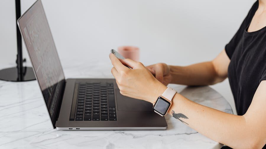 Person wearing a smartwatch and a black t-shirt, using a smartphone while sitting at a marble desk with a laptop open.