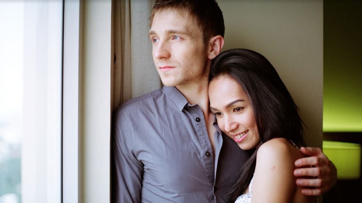 Cyril Mazur and Maki Gingoyon standing by a window, embracing each other with a warm expression.