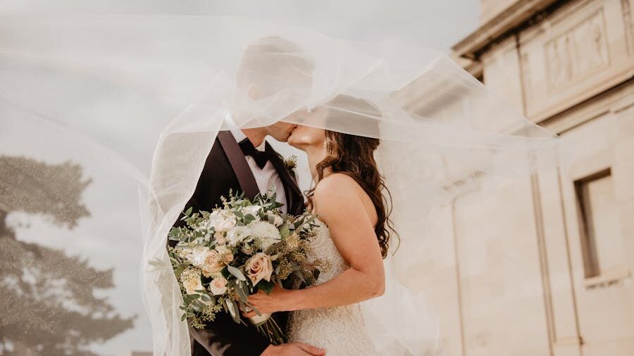 Bride and groom sharing a kiss under the bride’s veil, holding a bouquet of white and peach flowers.