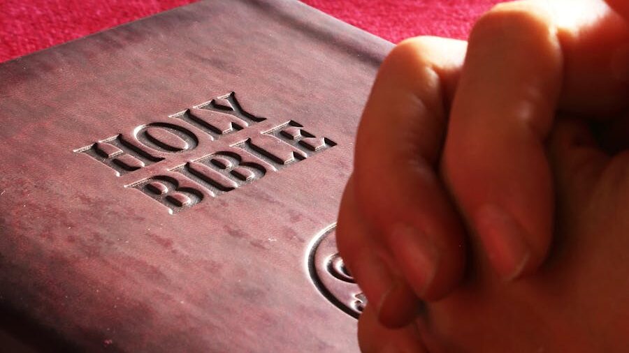 A person praying with hands clasped on top of a Holy Bible.