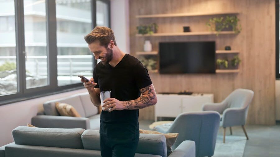 Smiling man in a black t-shirt, holding a glass and looking at his phone in a modern living room.