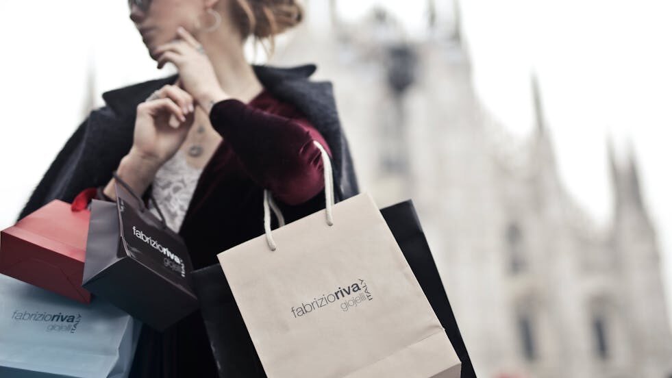 Stylish woman wearing sunglasses and a dark coat, carrying multiple shopping bags in an urban setting.