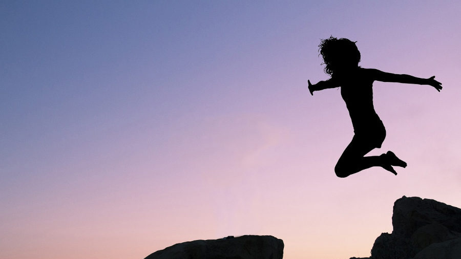 Silhouette of a person mid-jump between rocks at sunset, arms wide open against a purple and pink sky.