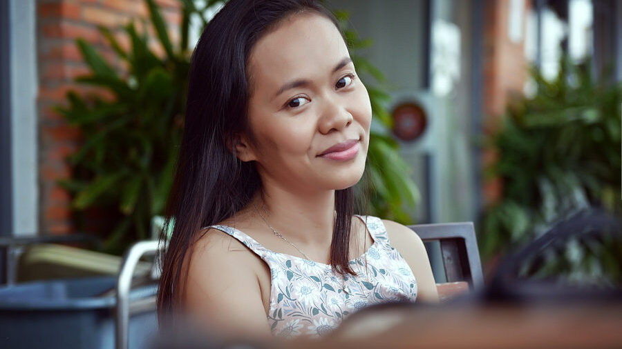 Woman sitting outdoors with a gentle smile, wearing a floral top, surrounded by plants and soft natural light.