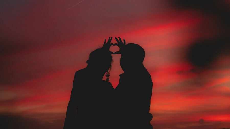 Two silhouetted figures making a heart shape with their hands in front of a vivid red-orange sunset sky.