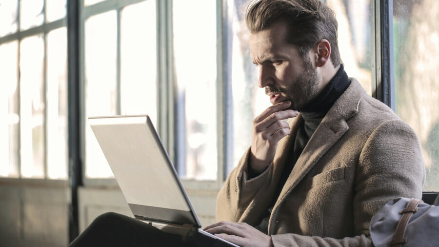 Man in a tan coat sits indoors, thoughtfully looking at his laptop, with natural light from large windows in the background.