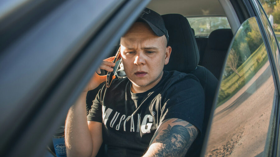 A man in a black shirt sits in the driver’s seat of a car, speaking on his phone with a serious and focused expression.