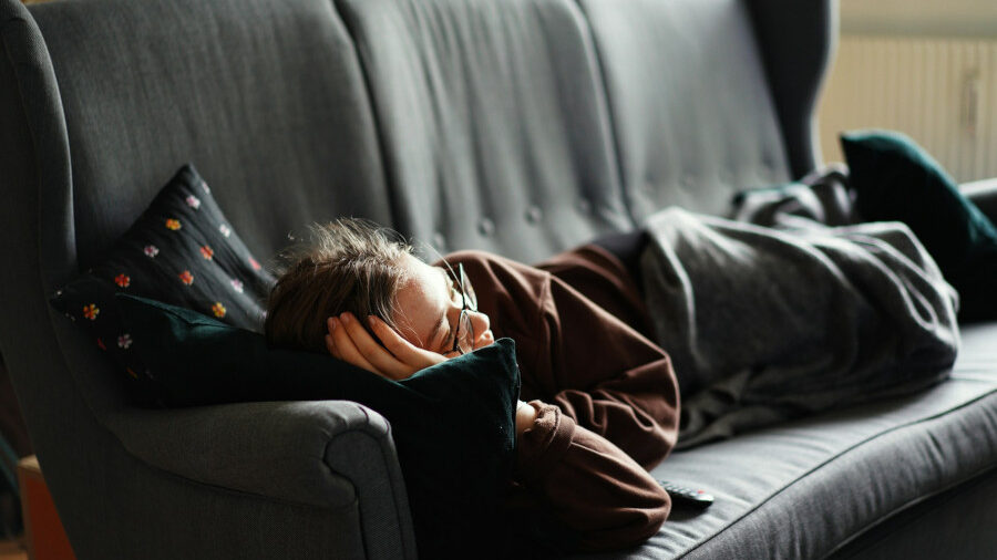 Person lying on a grey couch under a blanket, resting with head on a pillow in a quiet living room.