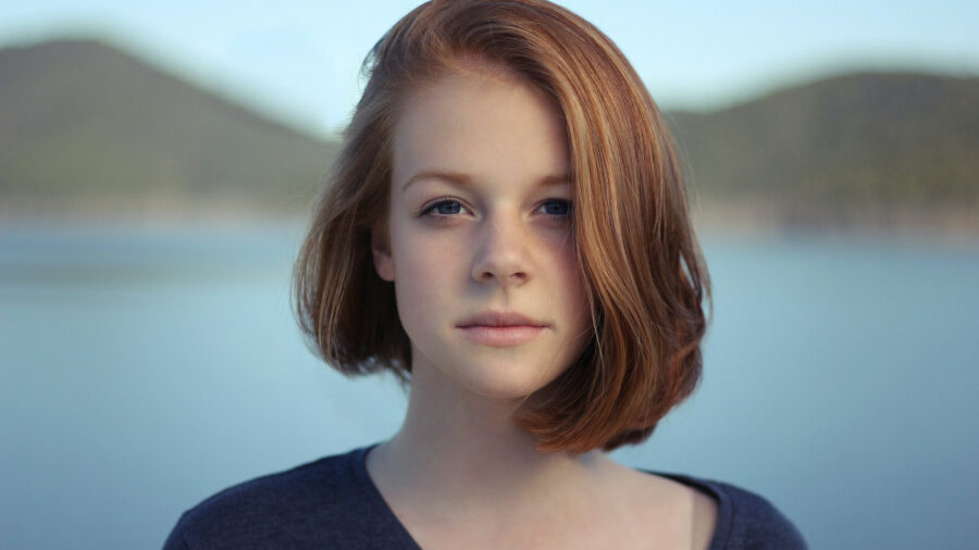 A young woman with auburn hair stands outdoors, looking calmly at the camera, with a lake and hills blurred in the background.