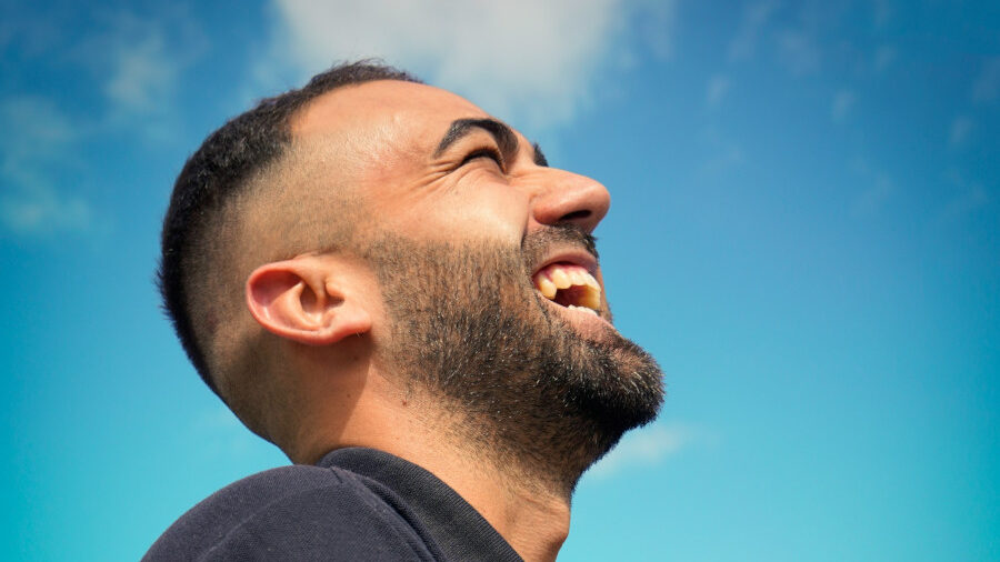 Close-up side profile of a bearded man laughing under a bright blue sky, showing an expression of joy and freedom.