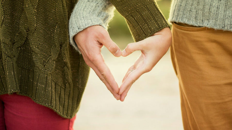 Two people wearing sweaters forming a heart shape with their hands, standing close in an outdoor sunlit setting.