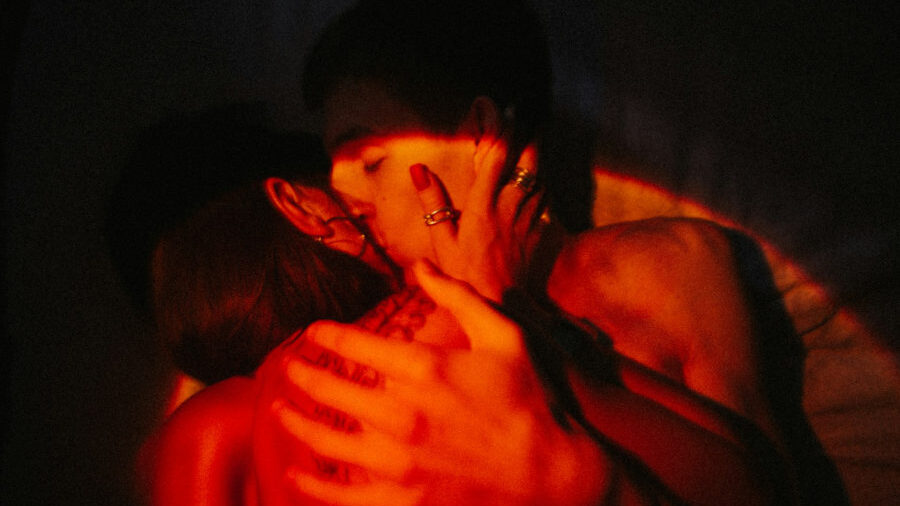 Two people kiss in a warm, shadowed environment, lit by soft red tones, their hands gently holding each other’s faces.